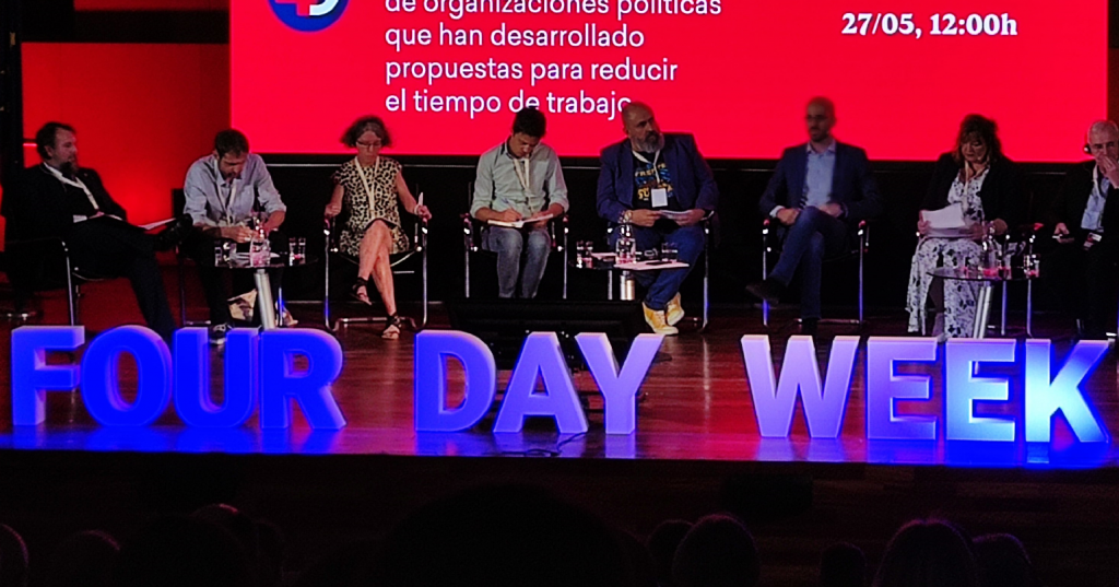 A group of people on a stage with a sign reading four day week