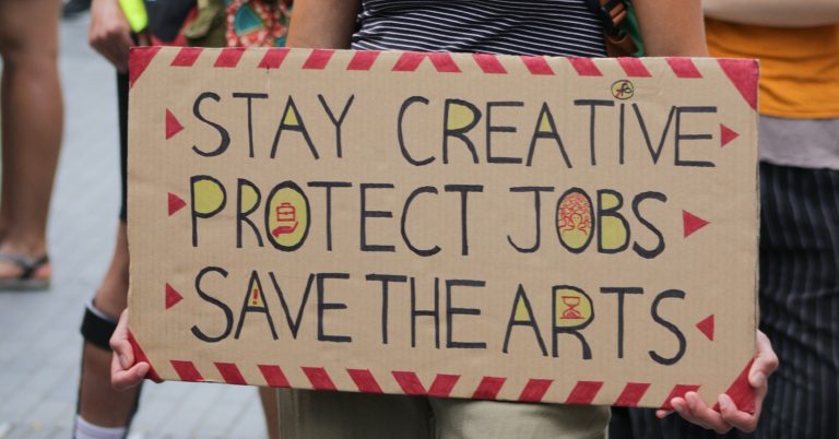 A placard reading "Stay creative, protect jobs, save the arts"