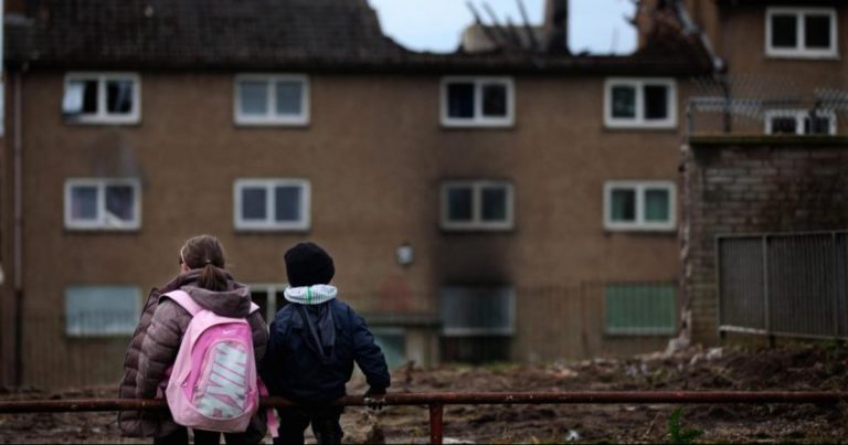 A photo of two children standing in front of run-down housing